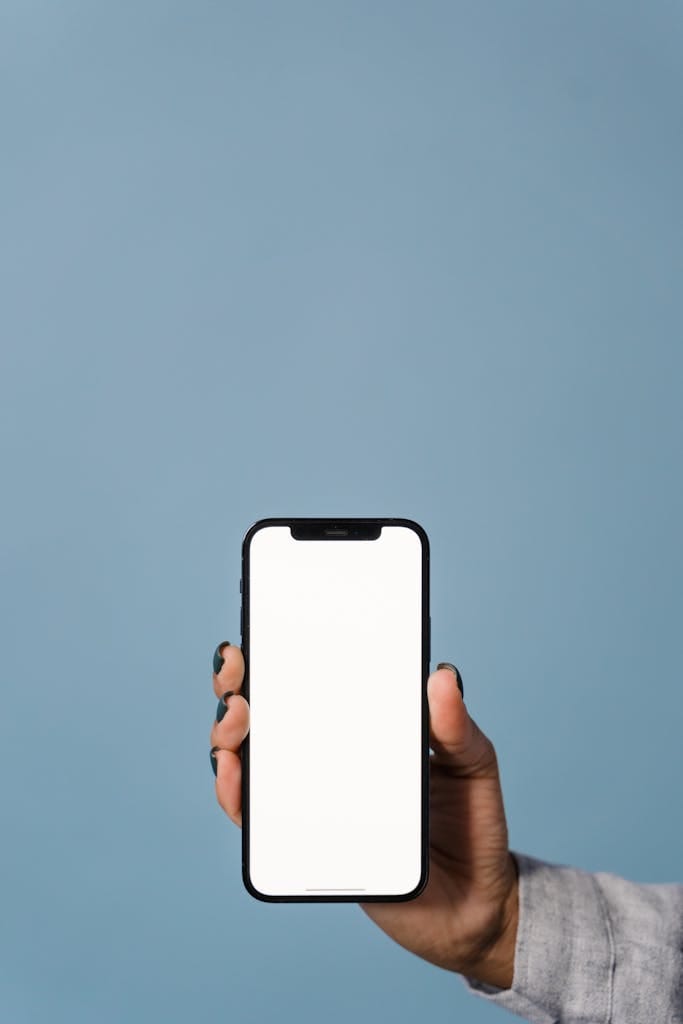 Primer plano de una mano sosteniendo un teléfono inteligente contra un fondo azul, mostrando una pantalla en blanco.