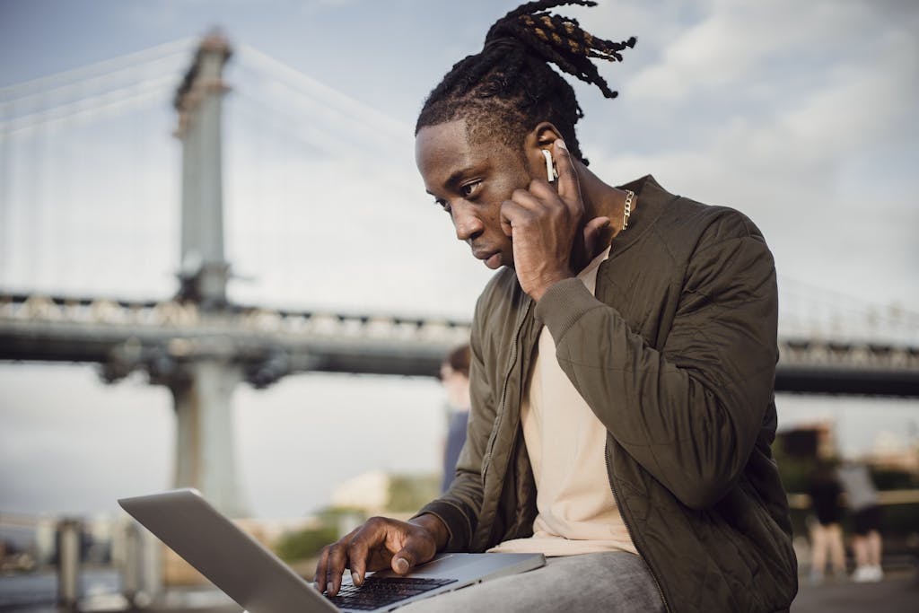 Ángulo bajo de un hombre negro concentrado con ropa casual usando una computadora portátil y auriculares mientras trabaja al aire libre en el parque