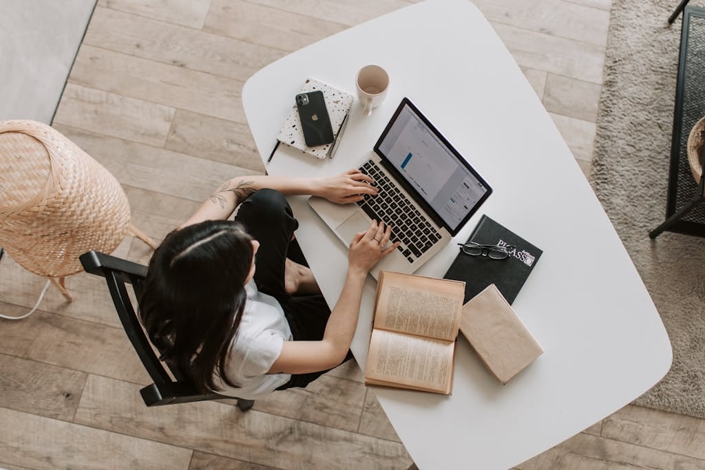 Vista aérea de una mujer usando una computadora portátil en el escritorio de una casa, rodeada de libros, un teléfono y una taza.