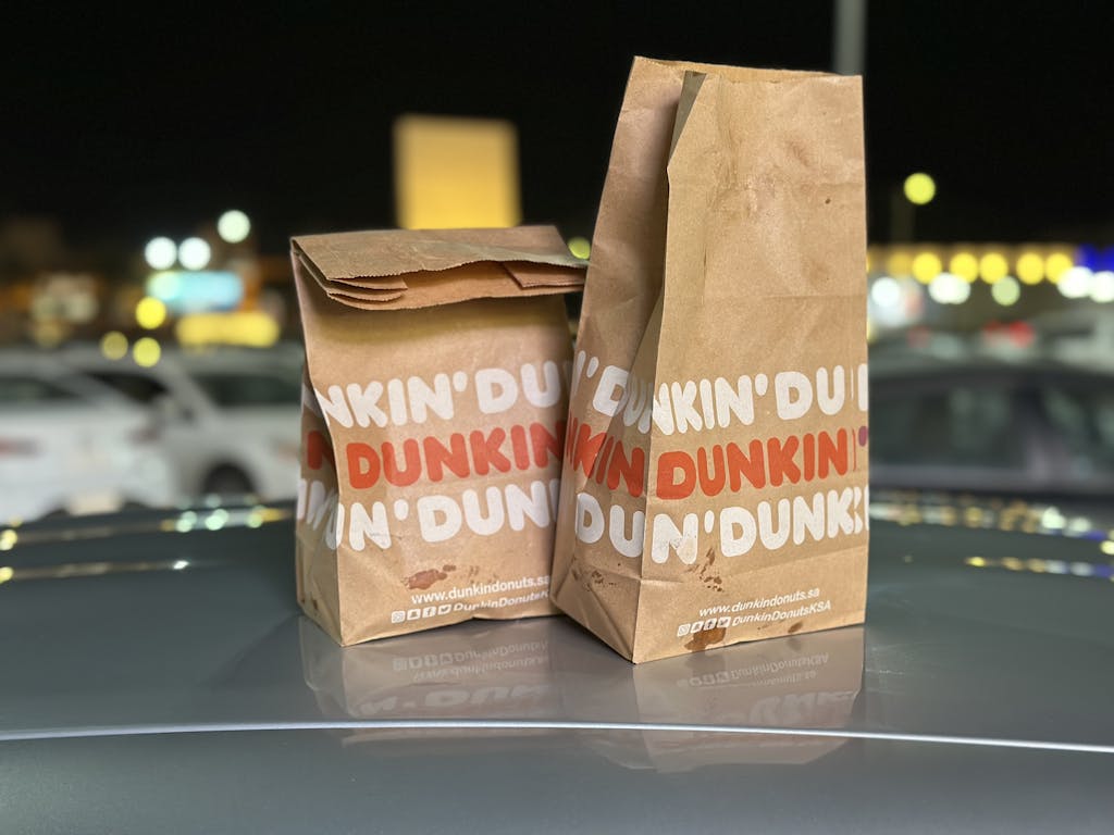 Dos bolsas de papel de Dunkin' Donuts colocadas en el capó de un coche con luces nocturnas borrosas de la ciudad en el fondo.